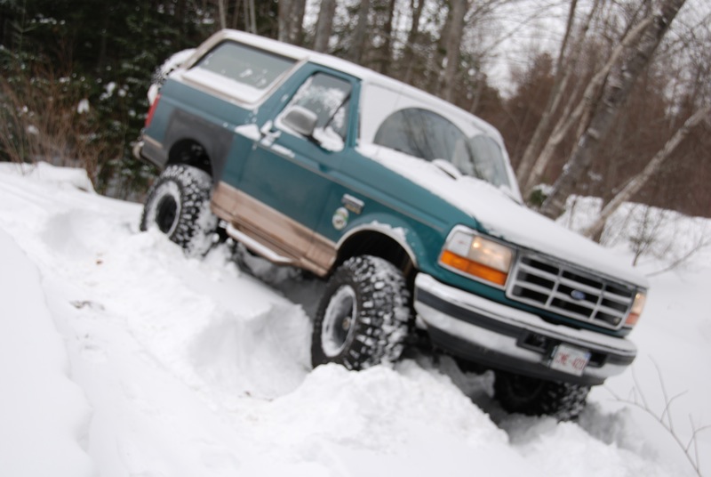Green and tan Ford Bronco driving through deep snow. License plate is visible
