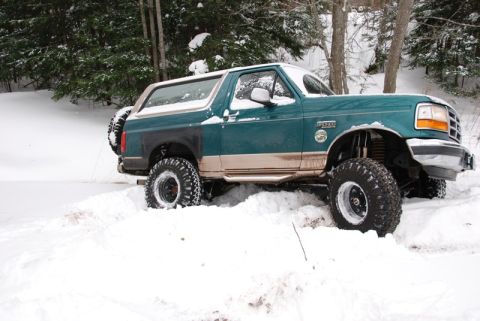 1996 Ford Bronco - A 1996 Ford Bronco navigates a snowy, wooded landscape, its large tires digging into the snow.