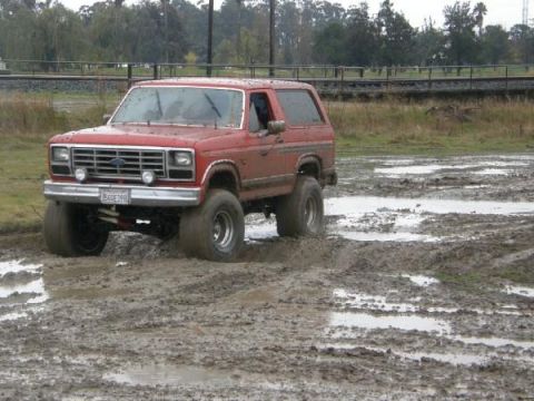 1986 Ford Bronco - Red 1986 Ford Bronco navigating a muddy terrain, tires deep in the mire.