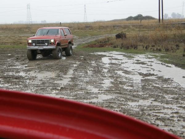 Red Ford Bronco with lifted suspension driving on a muddy track. The Bronco has a removable top