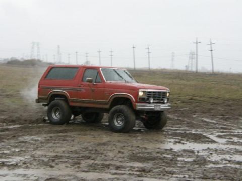 1986 Ford Bronco - A red SUV navigates a muddy terrain under an overcast sky, kicking up dirt.