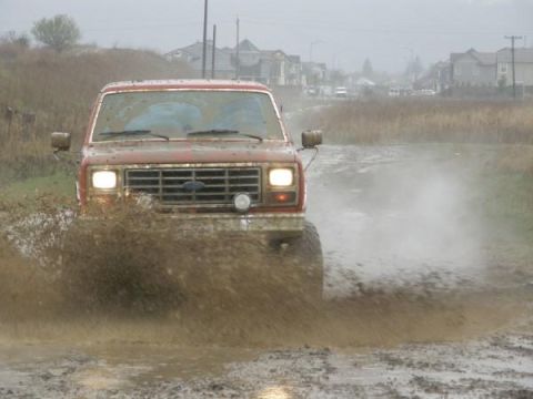 1986 Ford Bronco - A red Ford Bronco navigates a muddy track, kicking up water and dirt.