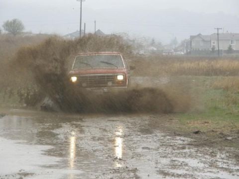 1986 Ford Bronco - A red 1980s Ford Bronco powers through a muddy puddle, creating a large splash.