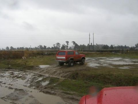 1986 Ford Bronco - A red SUV navigates a muddy terrain under an overcast sky. Another vehicle is partially visible in the foreground.