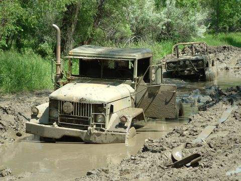 1992 Ford F-350 - Two military trucks are stuck in a muddy off-road trail with trees in the background.