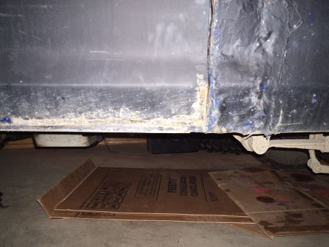 1979 Ford Bronco - Close-up of a vehicle's lower body panel and undercarriage, showing rust and some protective cardboard on the floor.