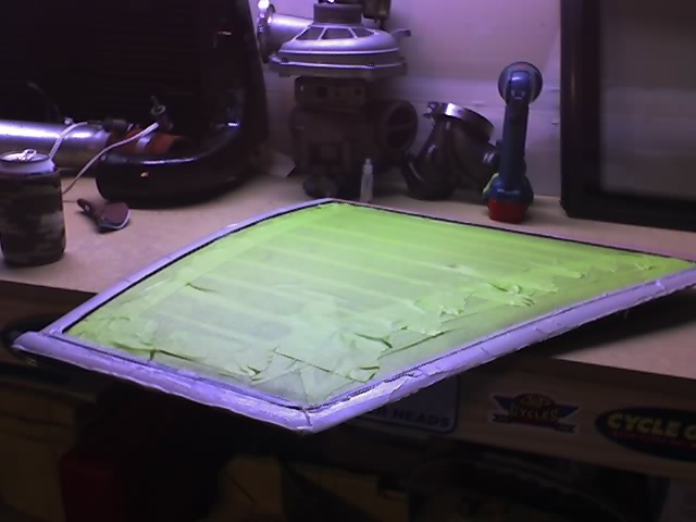 Car window pane covered in green masking tape, resting on a workbench with tools and parts in the background