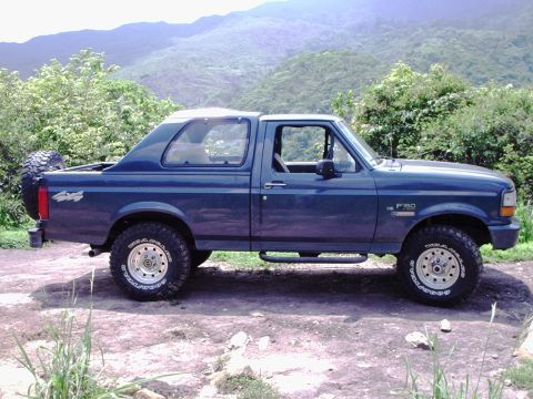 1995 Ford F-150 - A blue Ford Bronco is parked on a dirt road with a mountainous, tree-covered landscape in the background.