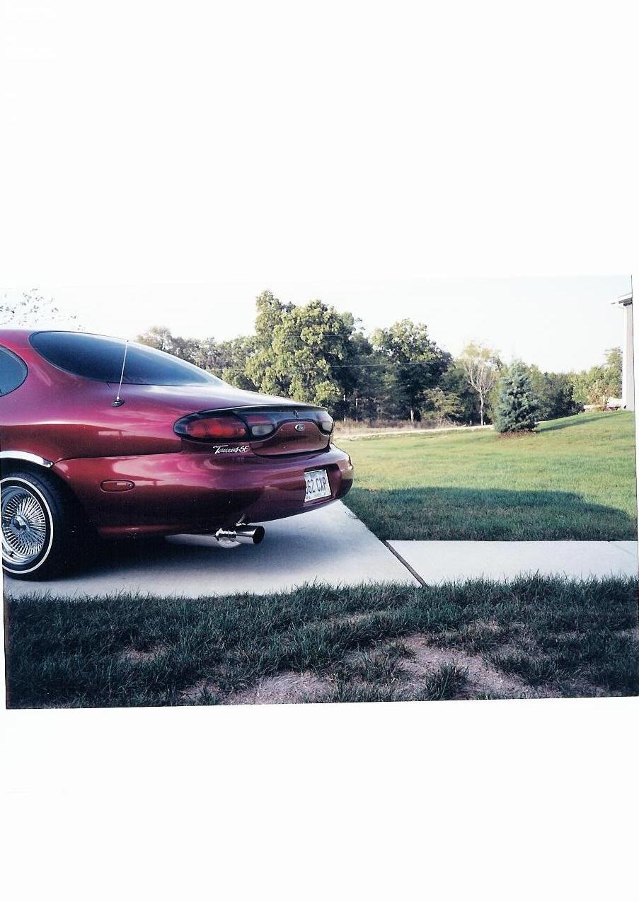 Rear of maroon Ford Taurus sedan with spoiler, wire wheels and chrome exhaust on a driveway.