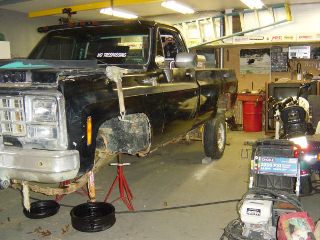 Front view of a black pickup truck on jack stands in a garage, undergoing repairs. A "NO TRESPASSING" sticker is on the windshield