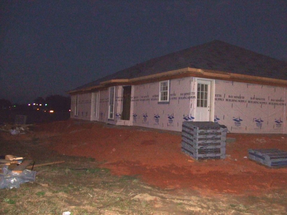 Exterior of a house under construction at dusk, with building wrap on the walls and a stack of materials in the foreground