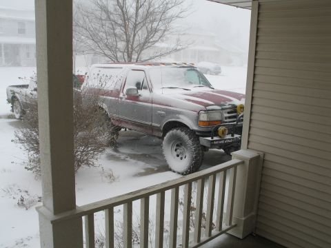 1995 Ford Bronco - Lifted two-tone red and silver 1990s Ford Bronco parked in a snowy driveway, shot from a covered porch through the railing during a snowfal…