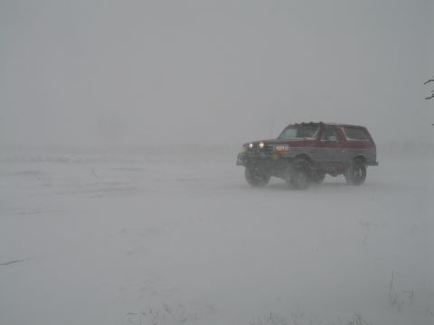 1995 Ford Bronco - A lifted red-and-silver two-tone Ford Bronco with oversized tires and roof clearance lights sits with headlights glowing in near-whiteout b…