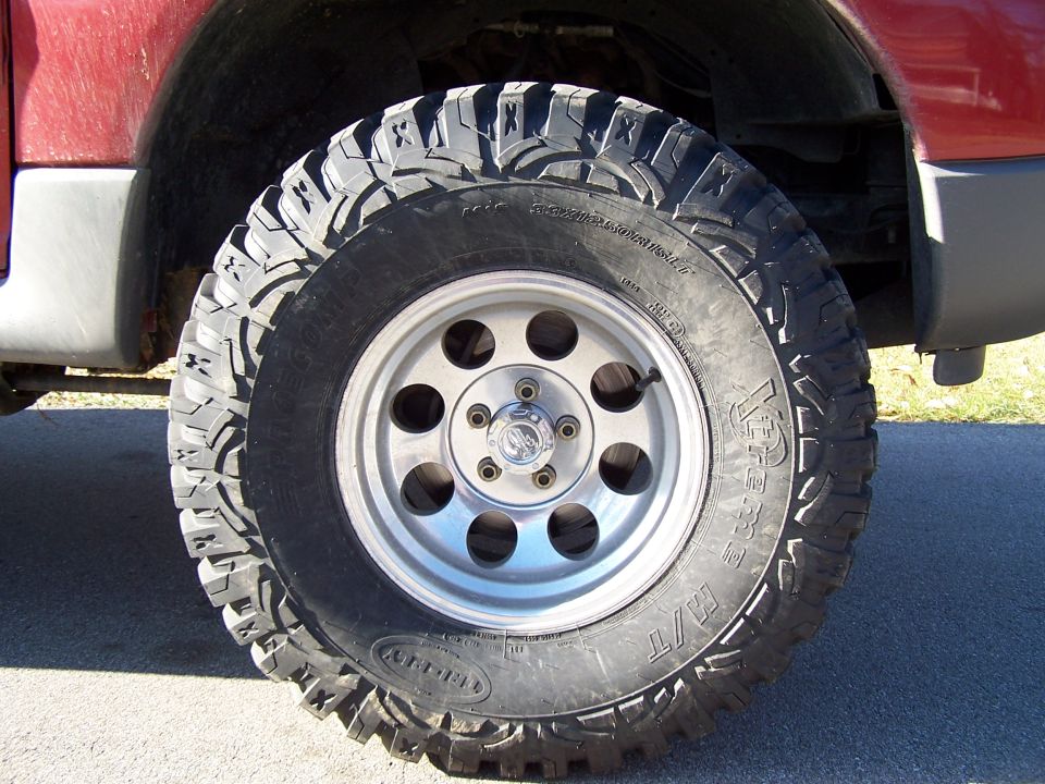 Close-up of a large off-road tire on a silver 5-spoke wheel, mounted on a red vehicle. Text on tire is partially visible