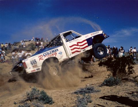 1985 Ford Bronco - A race truck soars through the air, leaving a trail of dust behind it against a clear blue sky.