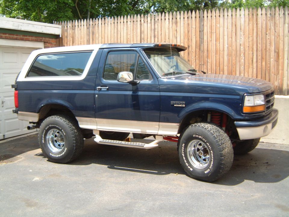 Lifted dark blue Ford Bronco SUV with white roof and BFGoodrich all-terrain tires, side view by a wooden fence
