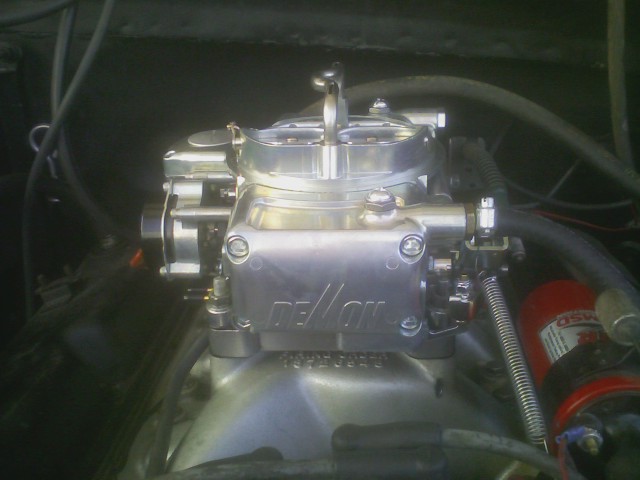 Close-up of a shiny Holley carburetor with "DEMON" visible on the side. Some hoses and engine components are in the background