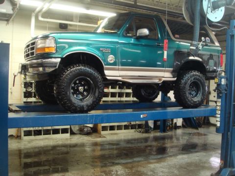 1996 Ford Bronco - A green Ford Bronco is raised on a hydraulic lift inside a garage, revealing its undercarriage and modified suspension.