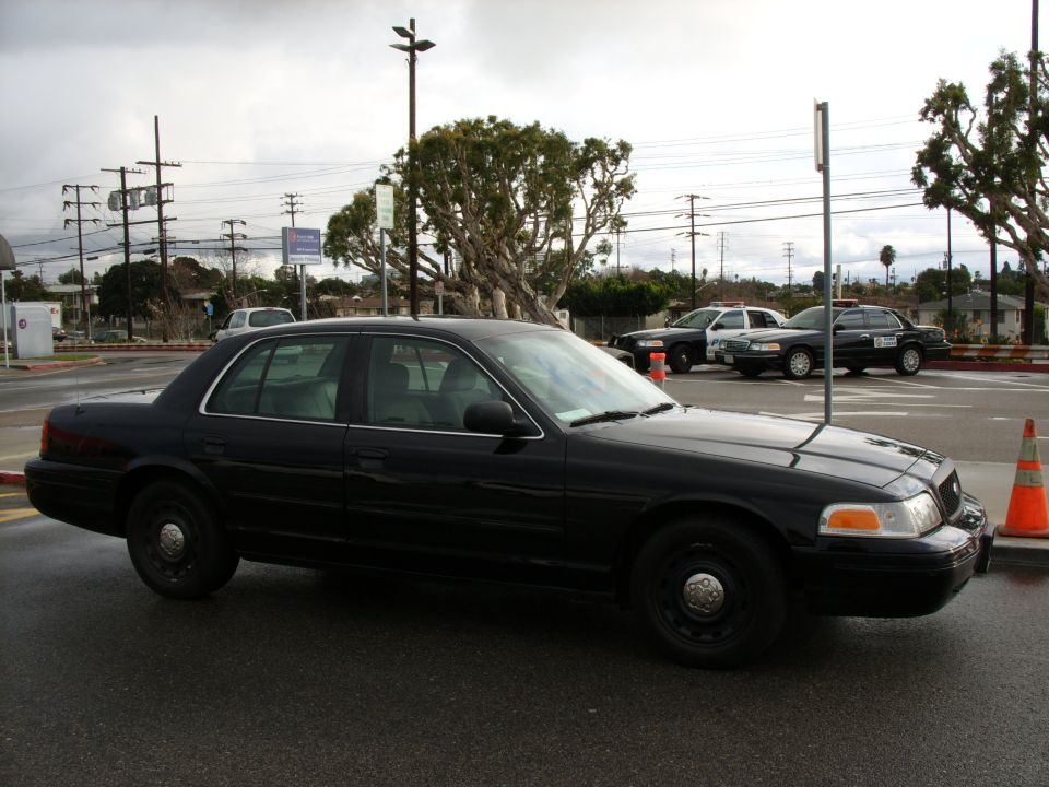 Black Ford Crown Victoria sedan with steel wheels beside police cruisers