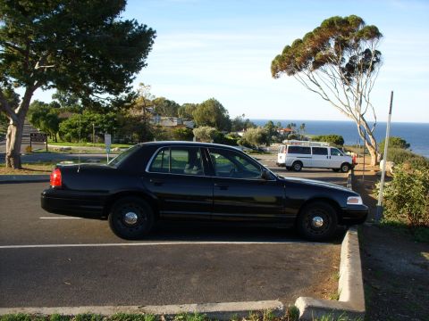 2005 Ford Crown Victoria - A black 2005 Ford Crown Victoria sedan is parked in a scenic spot overlooking the ocean, with a white van visible in the background.