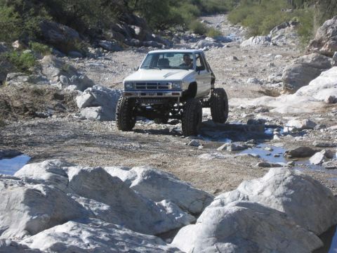 1987 Suzuki Samurai - This modified pickup truck navigates a challenging off-road terrain with large boulders and shallow water.