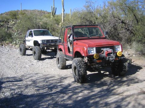 1987 Suzuki Samurai - Two modified off-road vehicles, a red Suzuki Samurai and a white Toyota pickup, are parked on a dirt road in a desert landscape.