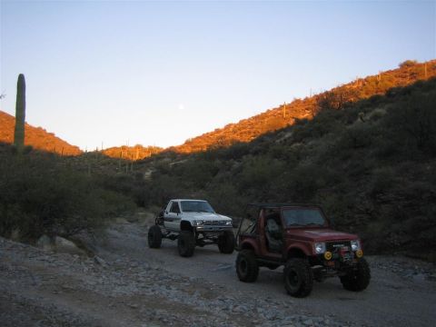 1987 Suzuki Samurai - Two modified off-road vehicles traverse a desert landscape at sunset, with a saguaro cactus visible.