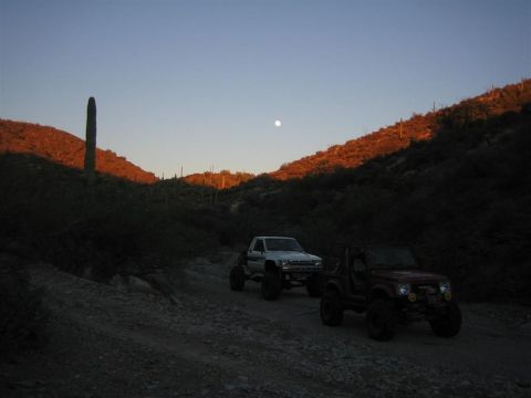1987 Suzuki Samurai - Two off-road vehicles, a red Suzuki Samurai and a white Toyota pickup, travel a dirt road through a desert landscape at dusk.