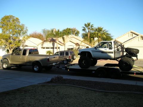 1986 Toyota Hilux - A gold pickup truck tows a trailer carrying a white pickup truck, with an SUV nearby.