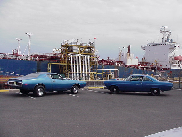 Two blue vintage cars parked at a dock with large ships and industrial piping behind them