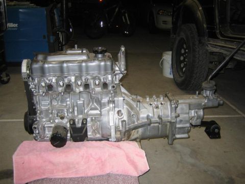 1987 Suzuki Samurai - A freshly cleaned four-cylinder engine bolted to a manual transmission and transfer case sits on a garage floor, photographed from the driv…