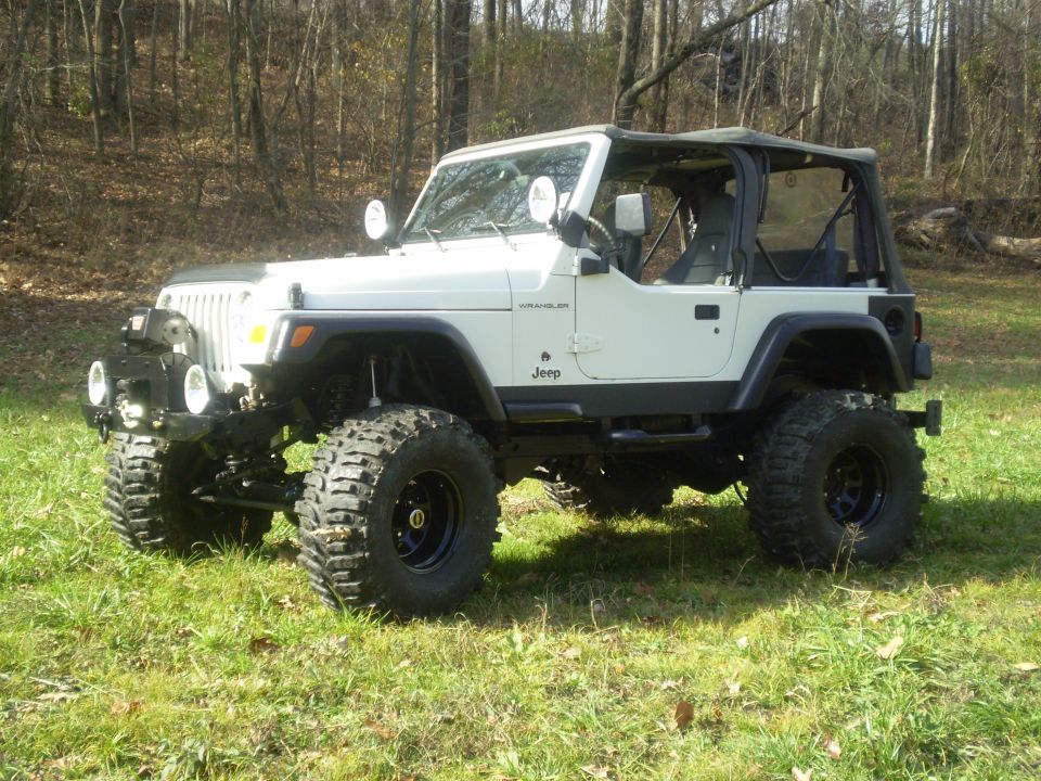 White Jeep Wrangler with aftermarket bumper and large tires parked on grassy hill
