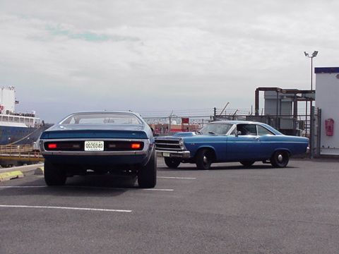 1966 Ford Fairlane - Two classic blue cars are parked in a lot near a large ship and industrial buildings.