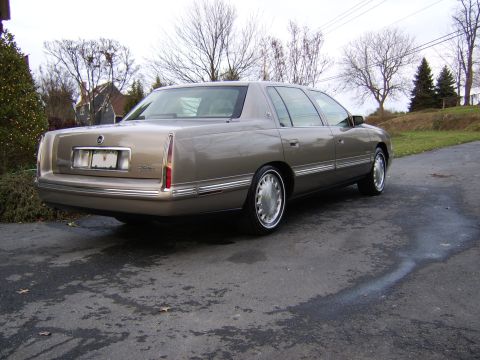 1996 Mercury Grand Marquis - A tan Cadillac sedan is parked on a wet asphalt driveway with trees and houses in the background.