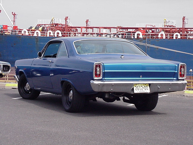 Blue 1960s Ford coupe rear 3/4 view on dock with red ship behind