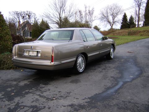 1996 Mercury Grand Marquis - A tan Cadillac sedan is viewed from the rear 3/4 angle on a wet driveway with trees and a house in the background.