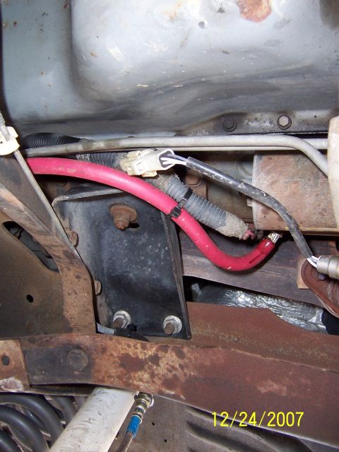 1994 Ford Bronco - Close-up view of the underbody wiring and frame of a vehicle, showing a red battery cable and various metal components.