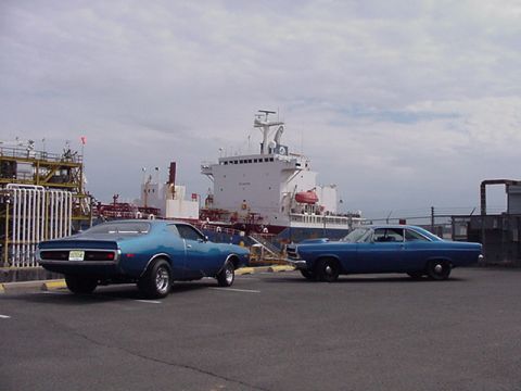 1966 Ford Fairlane - Two blue coupes are parked in front of a large ship and industrial complex under a cloudy sky.