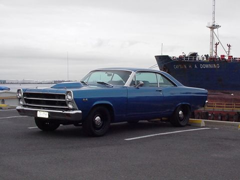 1966 Ford Fairlane - This blue 1966 Ford Fairlane coupe features a prominent hood scoop and is parked near a large ship.