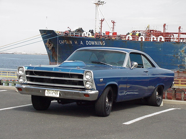 Blue 1966 Ford Fairlane coupe with hood scoop parked by a ship