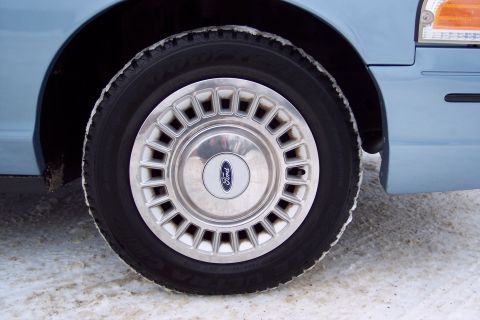 2001 Ford Crown Victoria - The front wheel of a light blue Ford Crown Victoria is shown in a snowy environment, with the Ford emblem clearly visible on the hubcap.