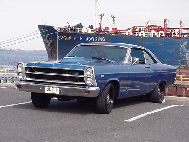 Blue coupe with a hood scoop parked in front of a large ship. License plate reads 2612-00