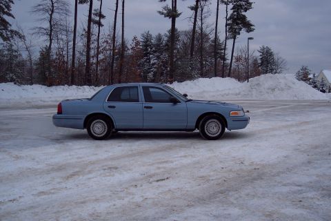 2001 Ford Crown Victoria - A light blue 2001 Ford Crown Victoria sits parked on a snow-covered lot under a cloudy sky.