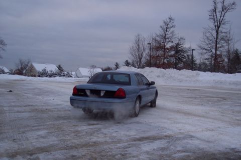 2001 Ford Crown Victoria - A blue sedan is seen from the rear, driving through a snow-covered area with smoke or snow spray coming from the tires.