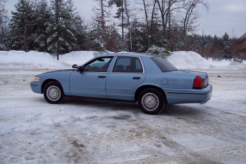 2001 Ford Crown Victoria - A blue 2001 Ford Crown Victoria sedan sits on a snow-covered surface with trees and snowbanks in the background.