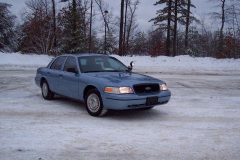 2001 Ford Crown Victoria - A blue 2001 Ford Crown Victoria sedan sits on a snow-covered lot under an overcast sky.