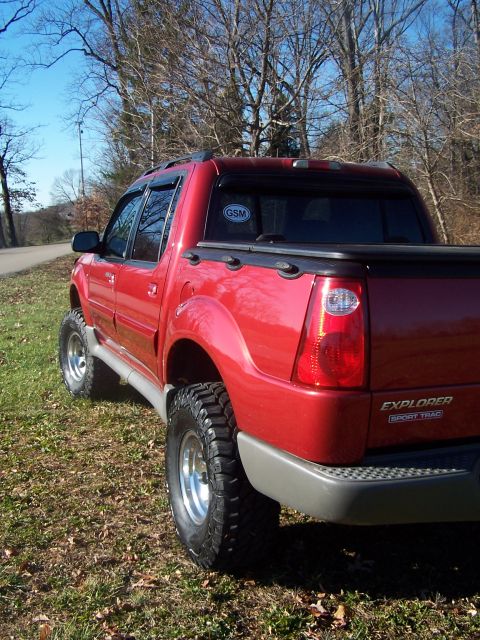2002 Ford Sport Trac - This red pickup truck has a lifted suspension and large off-road tires, parked on a grassy area with trees in the background.