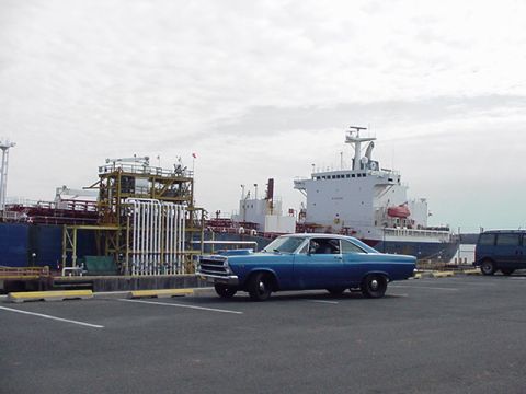 1966 Ford Fairlane - A blue 1966 Ford Fairlane is parked in a lot with a large cargo ship and industrial structures in the background.