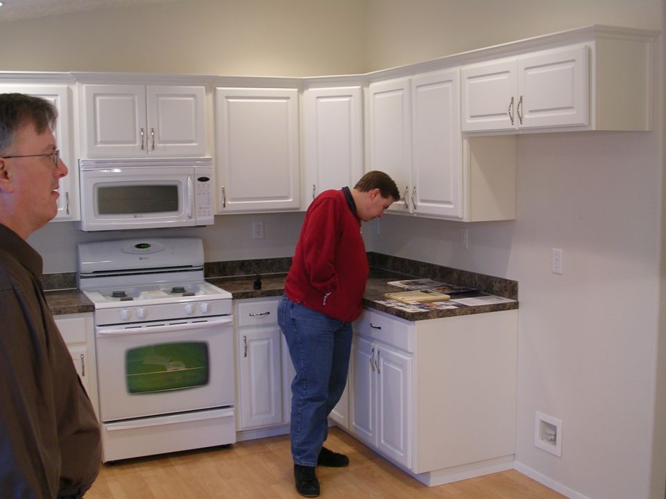 Two men in a kitchen with white cabinets, a white stove, and a microwave