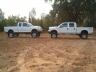 1990 Ford Bronco - Two white pickup trucks are parked on a dirt surface with trees in the background.